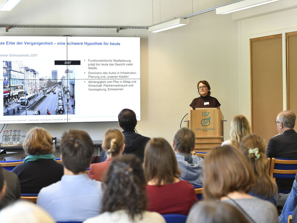 Eine Frau vor einer Leinwand, einen Vortrag haltend. Im Vordergrund Köpfe vom Publikum.